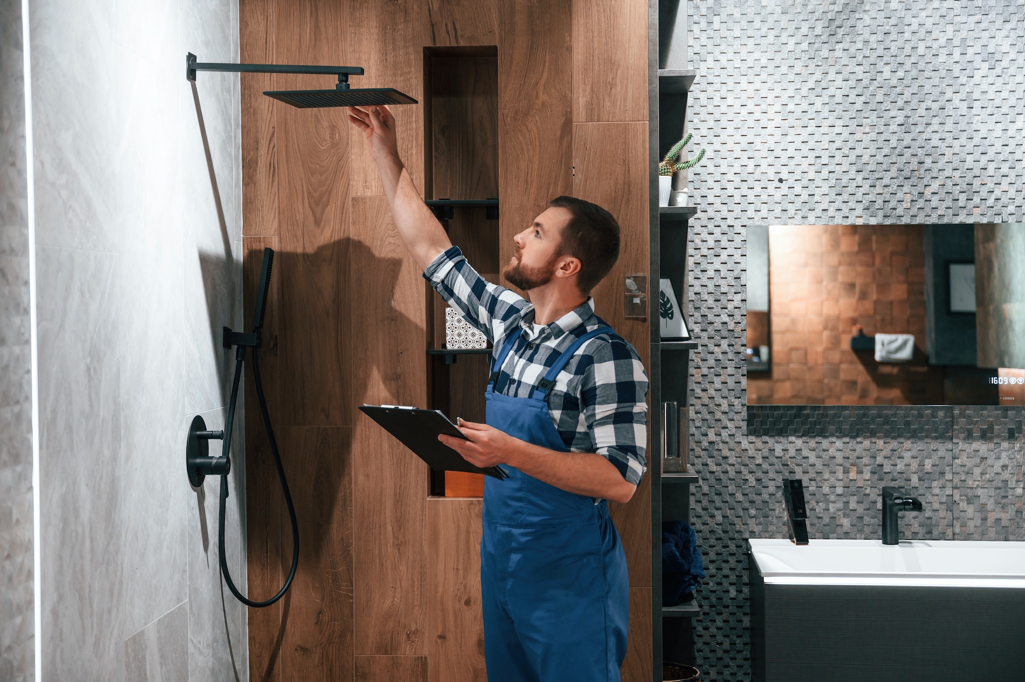 Repairs the shower faucet. Plumber in blue uniform is at work in the bathroom